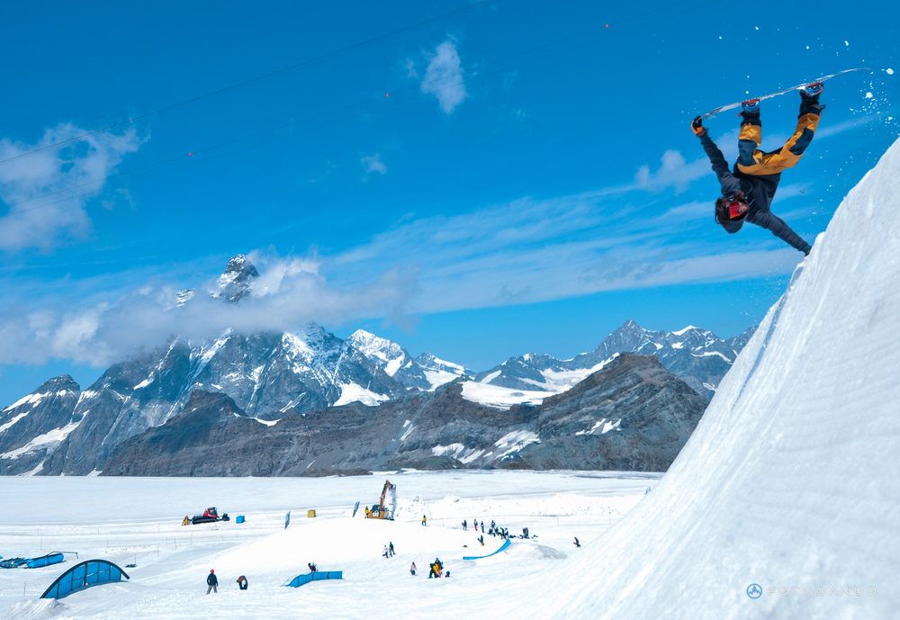 Snowboarder flying over a kicker with the snowy Alps behind, July 2021